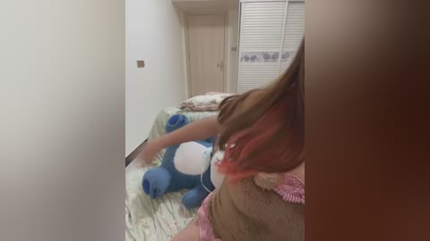 Media: Video of a young girl with red hair and a blue stuffed animal, sitting on a bed with a patterned blanket. The room features a white door and a closed white louvered closet.