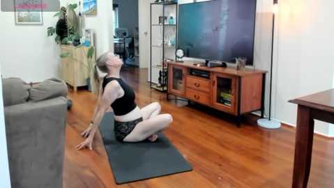 Media: A video of a woman with short, light hair doing yoga on a yoga mat in a living room with wooden floors, a TV, and a bookshelf.