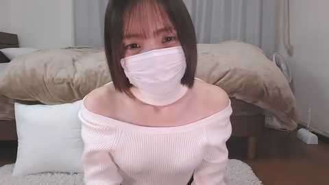 Media: A video of an East Asian woman with shoulder-length dark hair, wearing a white off-shoulder sweater, white mask, and kneeling on a shaggy white rug in a modern bedroom.