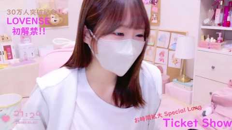 Media: A video of an East Asian woman with shoulder-length brown hair, wearing a white face mask, white top, and seated in a pink chair in a pastel-themed room with pink shelves and framed pictures. Text overlay in Japanese and English.