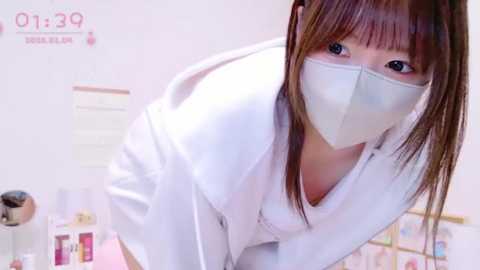 Media: A video of an Asian woman with long brown hair, wearing a white lab coat and a mask, leaning over a desk in a sterile laboratory setting.