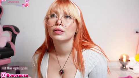 Media: Video of a fair-skinned woman with long, straight, orange hair and round glasses, wearing a low-cut white top, sitting in a gaming chair, surrounded by a lit room with a pink lamp and a gaming controller.