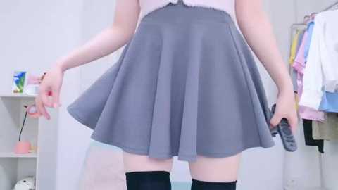 Media: Video of a young woman with fair skin wearing a high-waisted, gray pleated skirt and black thigh-high socks, standing in a brightly lit room with white walls and hanging clothes.