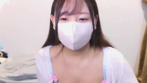 Media: Video of an East Asian woman with long dark hair, wearing a white surgical mask, a white robe, and pink flowers, indoors, blurry background.