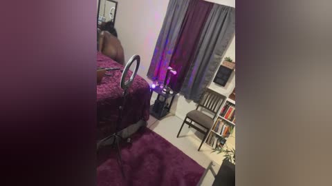 Media: Video of a dimly lit, small bedroom with dark purple bedding, a brown leather chair, and a desk with a computer monitor.