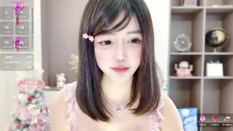 Media: Video of an East Asian woman with straight black hair, wearing pink lace top, smiling in a modern, brightly lit room with pink decor, shelves, and a globe.