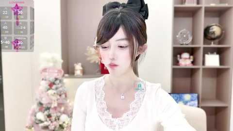 Media: Video of a young Asian woman with fair skin, black hair in a high bun, wearing a white lace-trimmed blouse, sitting indoors. Background includes a pink Christmas tree, bookshelves, and a TV screen.