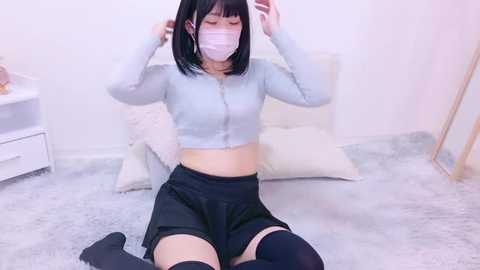 Media: Video of an East Asian woman with a slender build, short black hair, wearing a light blue crop top, black shorts, thigh-high socks, and a face mask, sitting on a soft, light-colored carpet in a simple room.