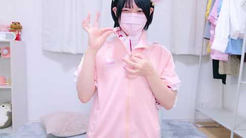 Media: Video of an Asian woman with short black hair, wearing a pink face mask, pink shirt, and black bow. She stands in a bright, clean room with white walls and a wardrobe.