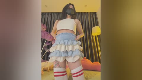 Media: Video of a young woman with a dark complexion, wearing a black mask, a white crop top, a ruffled blue skirt, and knee-high white socks with red stripes. She stands in a cozy, brightly lit bedroom with plush toys and a large lamp.