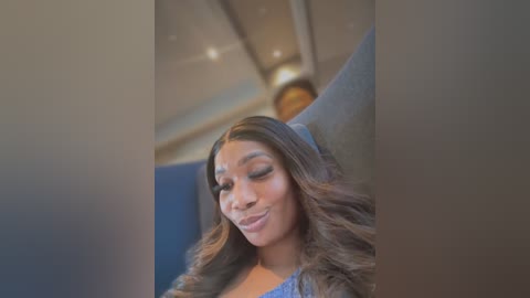 Media: Video of a young Black woman with long, wavy brown hair, wearing a blue top, smiling, in a modern, brightly lit airplane cabin with a clock on the ceiling.