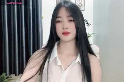 Media: Video of a young East Asian woman with long black hair, fair skin, and light makeup. She wears a white blouse, standing against a backdrop with a partially visible dark curtain and light walls.