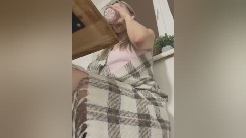 A candid video of a woman in a plaid blanket, drinking from a cup, leaning against a wooden table. Background includes a white-framed window and a small potted plant.