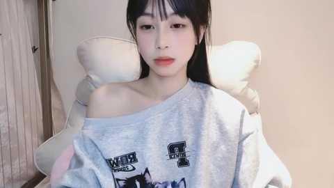 Media: Video of an East Asian woman with long black hair, fair skin, and small breasts, wearing a loose, off-the-shoulder grey sweatshirt, seated on a white leather chair in a beige room.