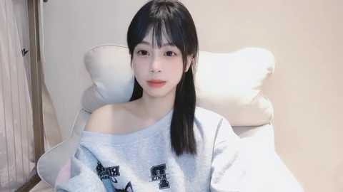 Media: Video of a young East Asian woman with straight black hair, fair skin, and a slim figure, wearing a loose, off-shoulder gray sweater. She sits on a white, cushioned chair in a softly lit room.