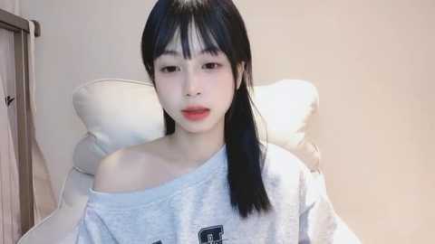 Media: Video of an East Asian woman with pale skin and long black hair, wearing a gray off-shoulder sweatshirt, seated on a white couch against a beige wall, looking serious.