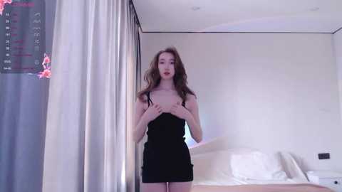 Media: A video of a slender, fair-skinned woman with long, wavy brown hair, wearing a black tank top and short skirt, standing in a modern, minimalist bedroom with white walls and a bed.