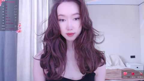 Video of a young Asian woman with long, wavy brown hair and fair skin, wearing a black top, standing in a bedroom with a white bed and curtains.