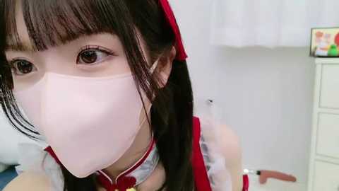 Media: Video of an East Asian woman with pale skin, wearing a white surgical mask, red hairband, and a white and red dress. Background shows a white curtain and a white dresser with colorful items.