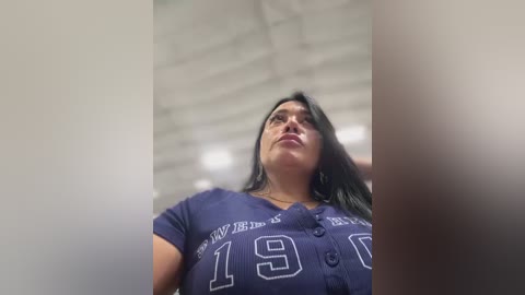 Media: Video of a middle-aged Latina woman with long black hair, wearing a navy blue \"49ers\" football jersey. She has a medium build and is looking upward. The background features an industrial ceiling with exposed beams.