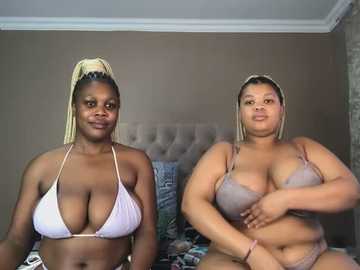 Media: Video of two Black women with dark skin, wearing bralettes, one with a low-cut purple bikini top, the other in a grey lace bralette. They sit on a bed in a beige room with patterned pillows.