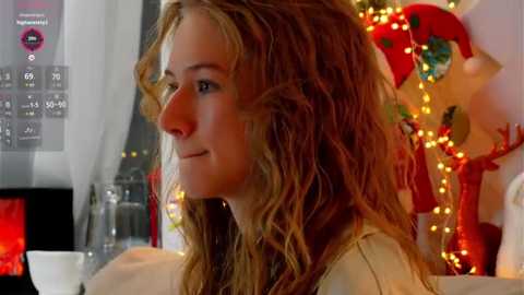 Media: Video of a fair-skinned woman with wavy, shoulder-length blonde hair, smiling, wearing a white top, in a cozy, festive room with Christmas lights, ornaments, and a fireplace.