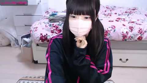 Media: Video of an East Asian woman with long black hair, wearing a face mask and black jacket, sitting on a bed with pink floral bedding in a cluttered bedroom.