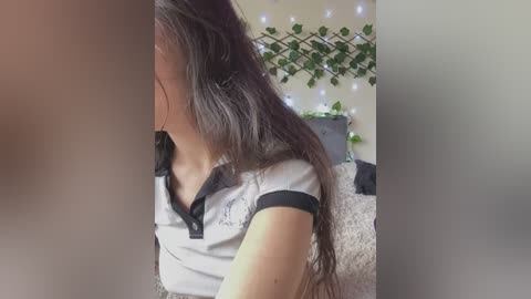 Media: Video of a woman with long, wavy dark hair, wearing a grey polo shirt, standing in a cozy room with green ivy and fairy lights on the wall.