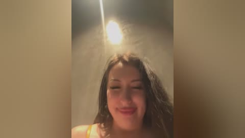 Media: A video of a smiling young woman with dark hair, wearing a yellow top, standing in a dimly lit room with a bright light source above her head, creating a halo effect.