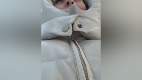 Media: Video of a person wearing a light beige puffy jacket with a zipper, partially obscured by the jacket's folds. The background is blurred, focusing on the jacket\u2019s texture and zipper detail.