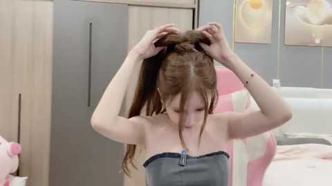 Media: Video of a young woman with light skin and long brown hair, sitting on a bed, using both hands to style her hair. The background features light-colored wooden furniture and floral wall art.