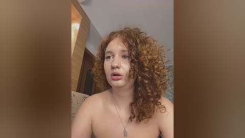 Media: Video of a young, topless, light-skinned woman with voluminous, curly, red hair, wearing a silver necklace. She stands indoors, with a beige and green patterned wall and wooden decor in the background.