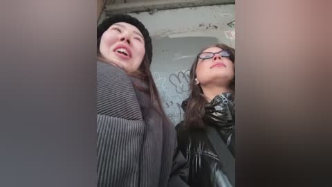 Media: Video of two women, one with long hair in a black beanie, the other with glasses and dark hair, laughing in a graffiti-covered, dimly lit room.