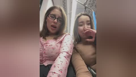 Media: Video of two young women in a subway car, one wearing a pink floral blouse and glasses, the other with a brown top, both with flushed faces, capturing a moment of intimate exchange.