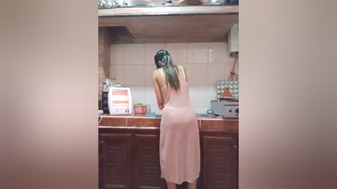 Media: Video of an Asian woman in a pink nightgown standing in a cluttered kitchen, facing away from the camera.