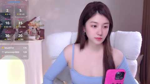 Media: Video of a young East Asian woman with long brown hair, wearing a light blue off-shoulder top, sitting on a white couch, holding a pink phone, with a digital weather overlay showing 10\u00b0C and cloudy skies.