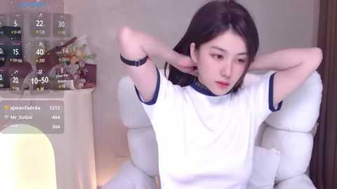 Media: A video of a young Asian woman with long black hair, wearing a white t-shirt, adjusting her hair in a modern living room with a white couch and a bookshelf.