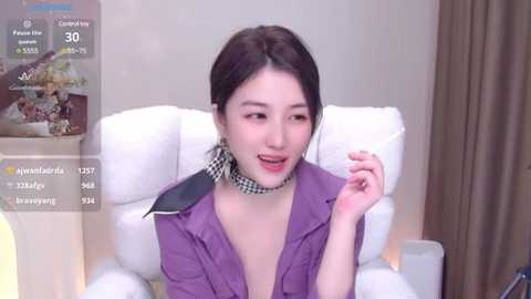 Media: Video of an Asian woman with fair skin, wearing a purple jacket, black-and-white patterned choker, and dark hair in a bun. She sits on a white chair in a room with beige walls.