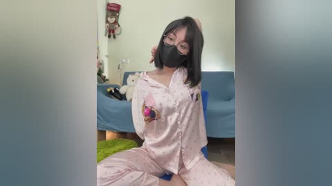 Media: Video of an Asian woman in pink pajamas with a mask, sitting on a green rug in a cozy, dimly lit room with a blue couch, Christmas decorations, and stuffed toys.