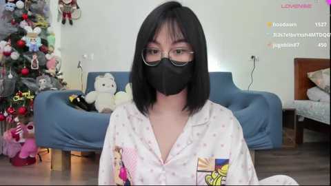 Media: Video of an Asian woman with shoulder-length black hair, wearing glasses and a black mask, in a pastel-patterned pajama top. Background includes a decorated Christmas tree, stuffed toys, and a blue sofa.