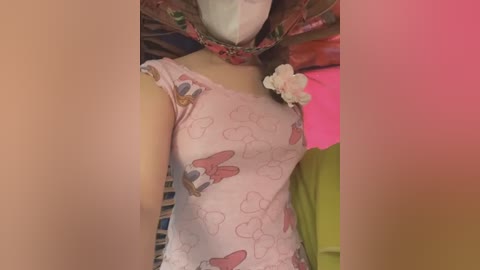 Media: Video of a person wearing a pink, butterfly-patterned shirt, a floral headscarf, and a white mask, standing in a room with pink and green decor.