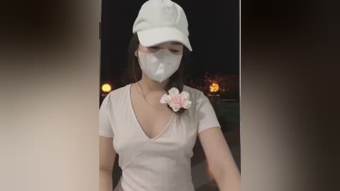 Media: Video of a young woman with light skin, wearing a white cap, mask, and short-sleeved shirt with a pink flower brooch. She stands outdoors at night, with blurred background lights.