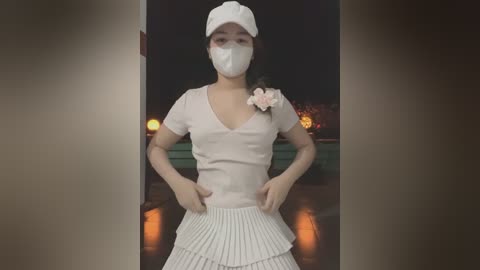 Media: Video of an Asian woman with a light complexion, wearing a white cap, mask, and a beige V-neck top with a pink flower brooch. She stands in a doorway with a dark background and a lit pumpkin on a green table behind her.