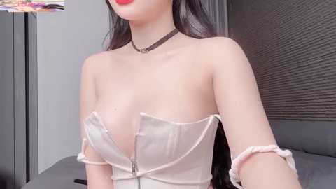 Video of a fair-skinned woman with long black hair, wearing a strapless, white corset with a visible zipper, black choker, and pink wristbands, posing indoors with a grey wall and blinds in the background.