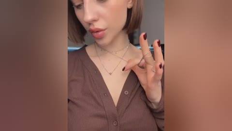 Media: A video of a fair-skinned woman with a bob haircut, wearing a brown cardigan, silver necklaces, and red-painted nails, making a peace sign.