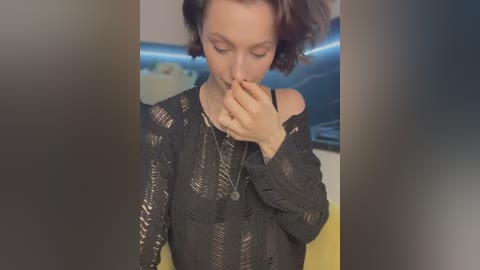 Media: A video of a fair-skinned woman with short, curly brown hair, wearing a black, off-shoulder knit sweater. She's covering her mouth with her hand, looking down. Background is blurred, featuring a modern kitchen with dark cabinets and a countertop.