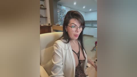 Media: A video captures a middle-aged woman with glasses, fair skin, and long black hair, wearing a white blazer over a revealing black lace bra, seated in a modern kitchen.