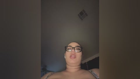 A video of a plus-sized woman with fair skin, wearing black-framed glasses, and a leopard print bra, making a pouty face, standing in a dimly lit room with a small vent on the ceiling.