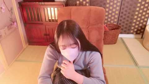 Media: A video of a young Asian girl with long black hair, wearing a gray hoodie and a face mask, sitting on a tatami mat in a traditional Japanese room with wooden furniture and a wicker basket.