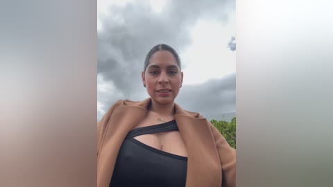 Video of a woman with light brown skin, wearing a tan coat over a black one-shoulder top, standing in front of a cloudy sky with green trees in the background.